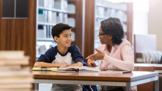 A smiling middle school age boy sits at a table with a mature female tutor in his school library. He listens as she gestures and explains the homework.