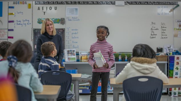 An African American elementary school students stands in front of her classroom and is preparing to talk about her project to her classmates.