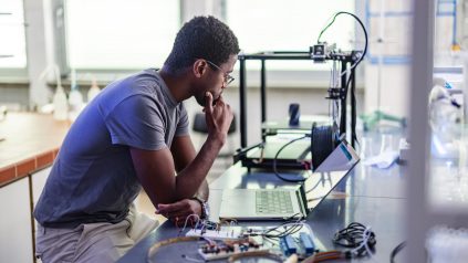 Young African-American technician sitting in the chair, thinking and repairing his 3D printer in the laboratory.