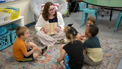 Female teacher sits with three elementary aged students and reads a book